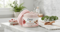A jar of Dove Exfoliating Body Scrub with pomegranate and shea butter, shown open with pink scrub visible, styled on a marble countertop with a bowl of scrub, greenery, and a towel in a bright bathroom setting.