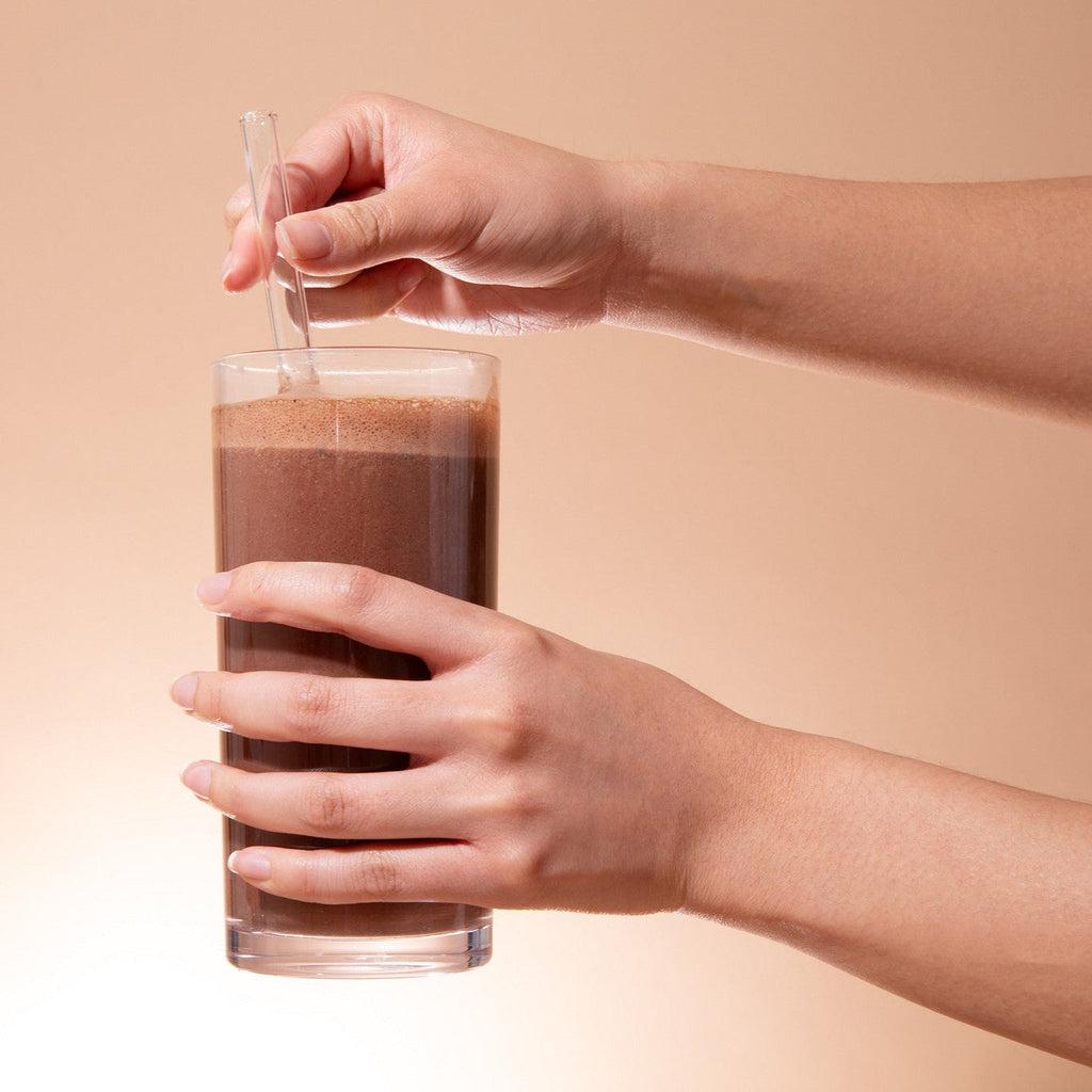 Person holding a glass of chocolate milk with a straw against a beige background