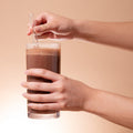 Person holding a glass of chocolate milk with a straw against a beige background