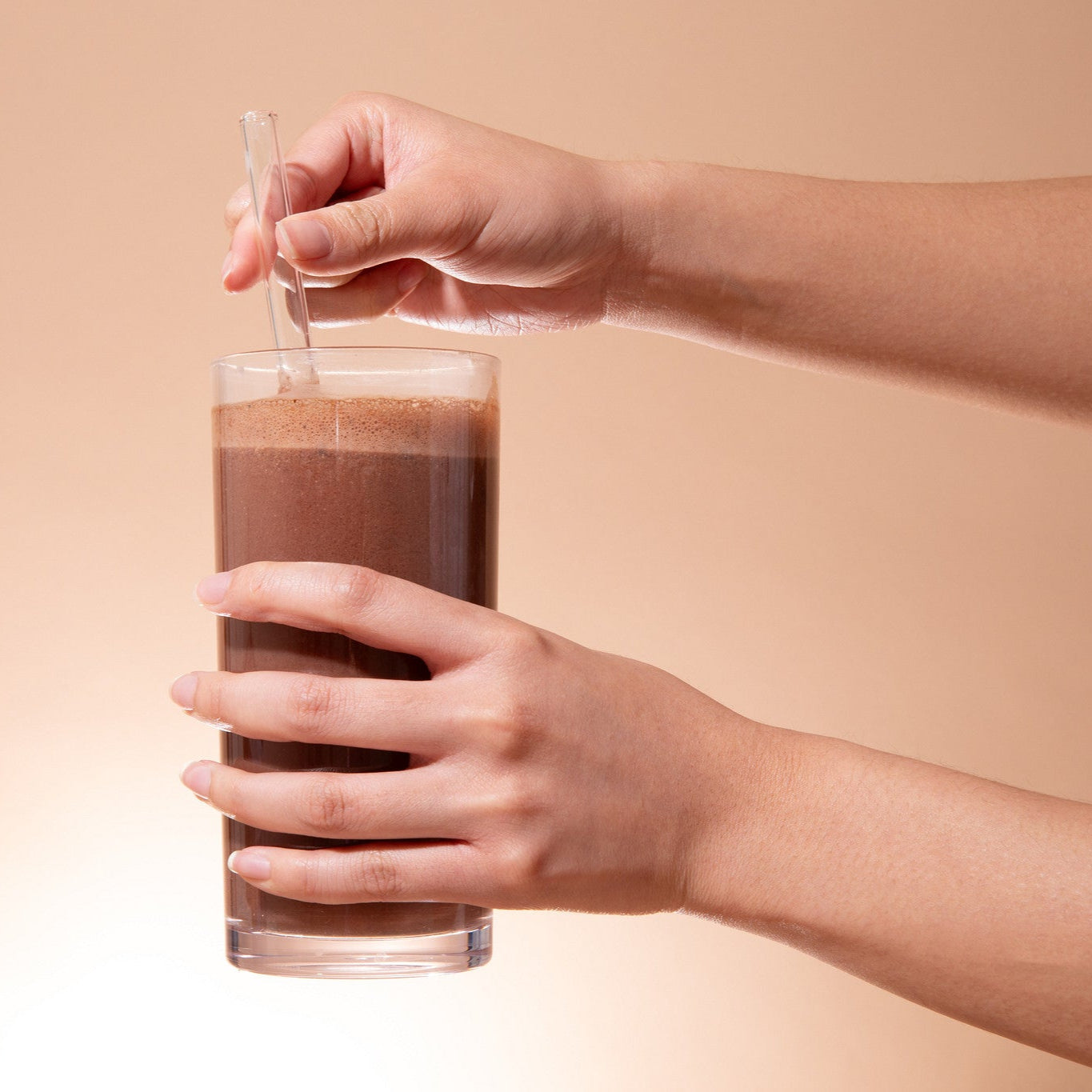 Person holding a glass of chocolate milk with a straw against a beige background