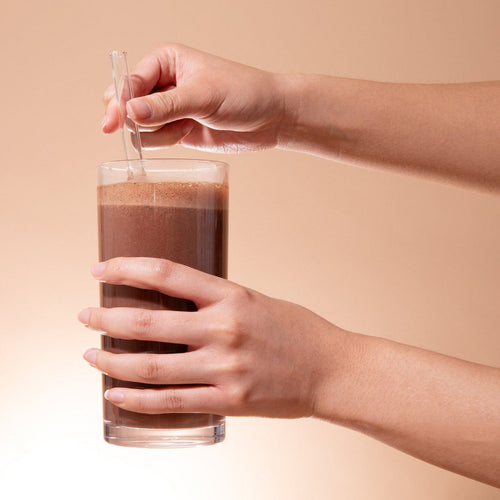 Person holding a glass of chocolate milk with a straw against a beige background